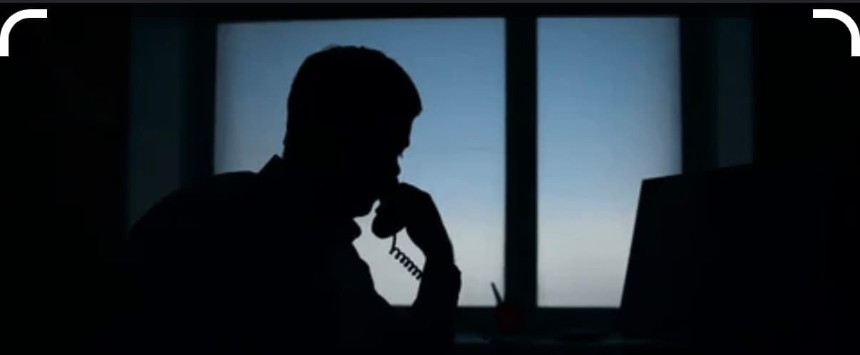 Silhouette of a person talking on a corded telephone at a desk by a window.